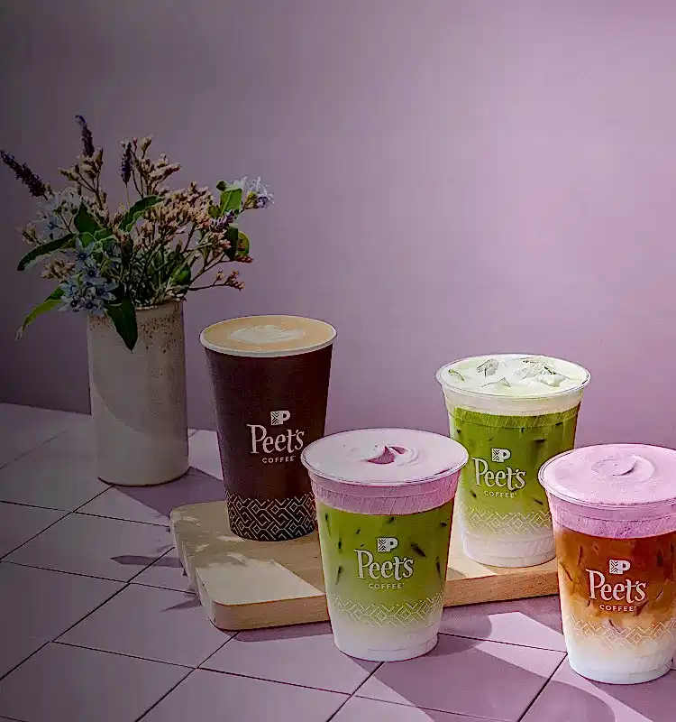 Peet's Coffee cups on a wooden board with a vase of flowers on a tiled floor.